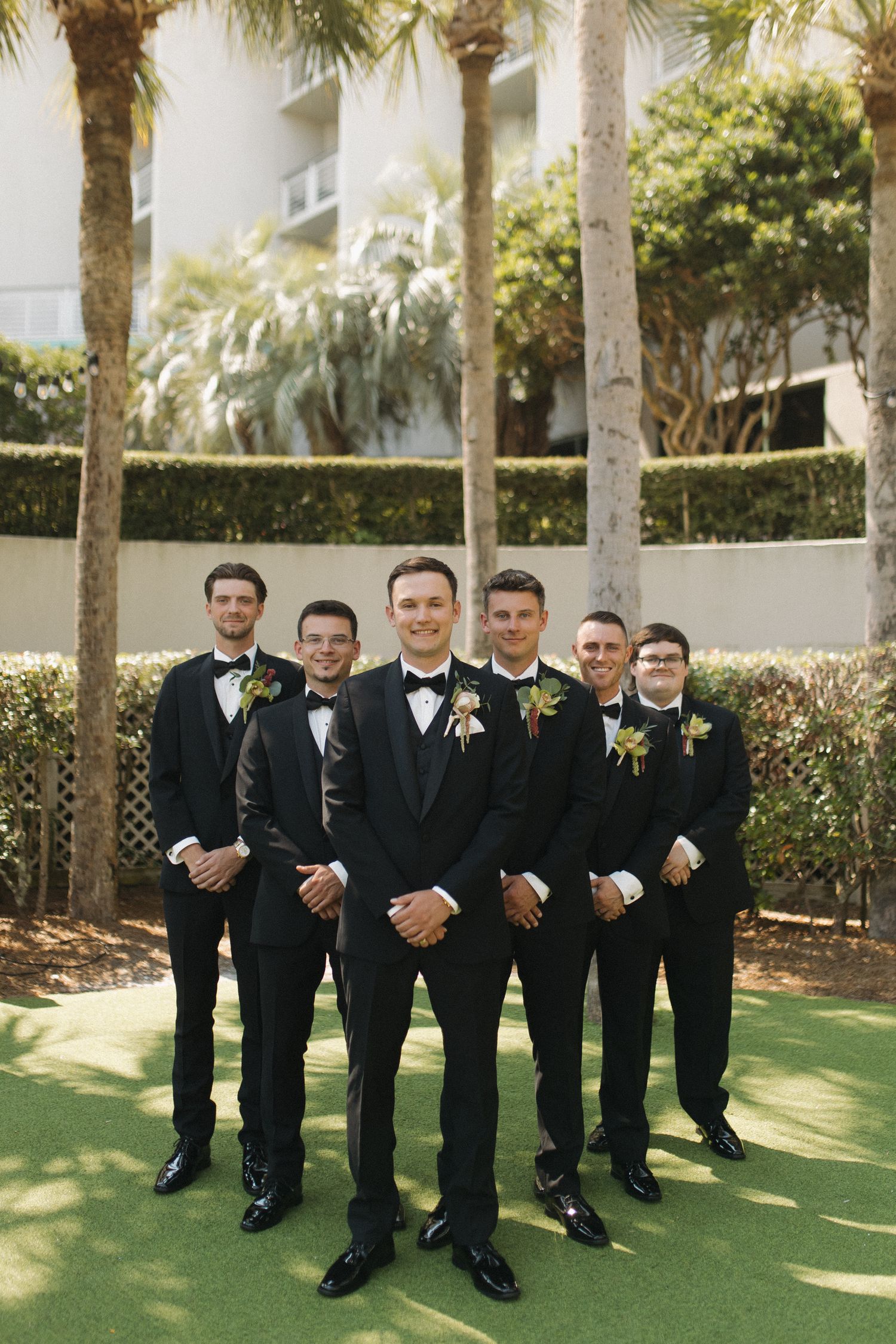 Groom and groomsmen in black tuxedos with boutonnieres pose outdoors near palm trees and hedges.