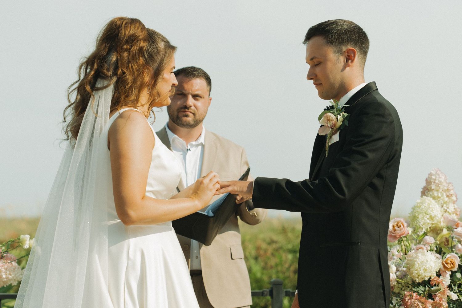 Bride and groom exchange vows outdoors with officiant, bride in white gown and veil, groom in black suit with floral boutonniere.