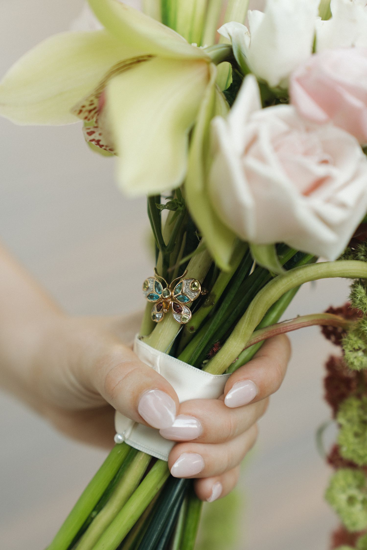 Hand holding a bridal bouquet with pink roses, white orchids, and a decorative butterfly brooch, wrapped in white ribbon.