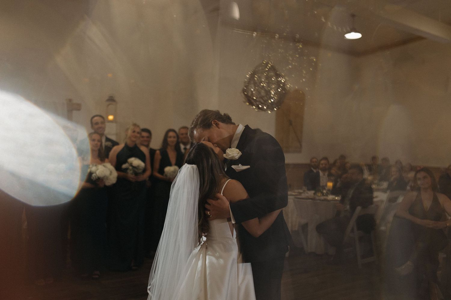 Bride and groom share a romantic first dance kiss at their wedding reception with bridesmaids and guests watching.
