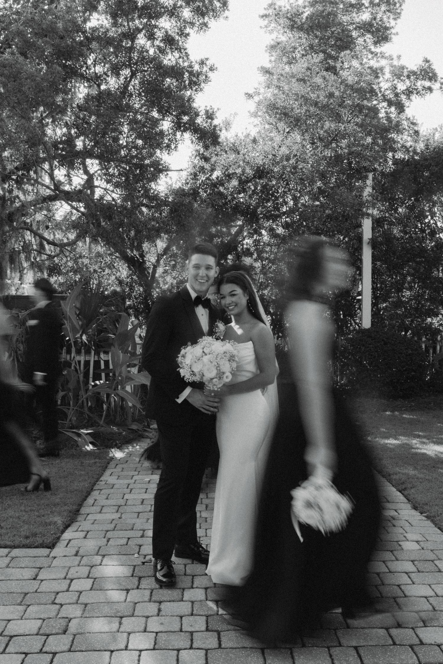 Bride and groom pose together holding bouquet on brick path in black and white wedding photo.