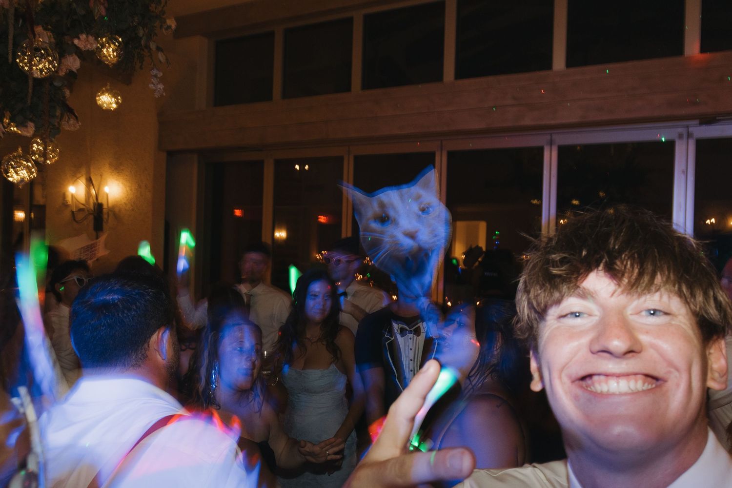 Smiling man takes selfie at lively night party with glowing neon sticks and dancing crowd indoors.