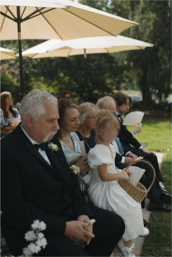 Wedding guests seated outdoors under cream umbrellas, including a flower girl in white dress holding a wicker basket.