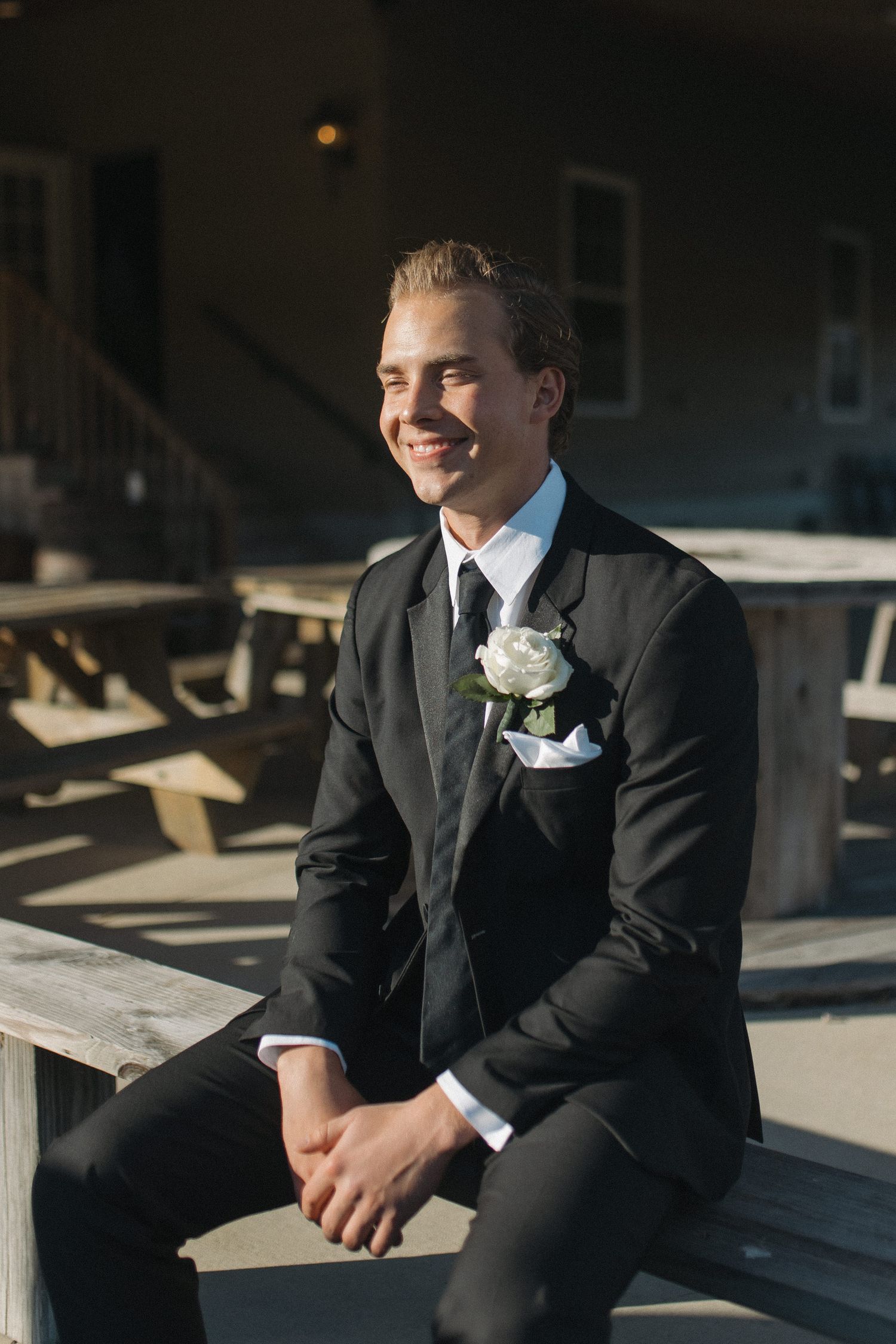 Smiling groom in black suit with white boutonniere sits on wooden bench outdoors at wedding venue.