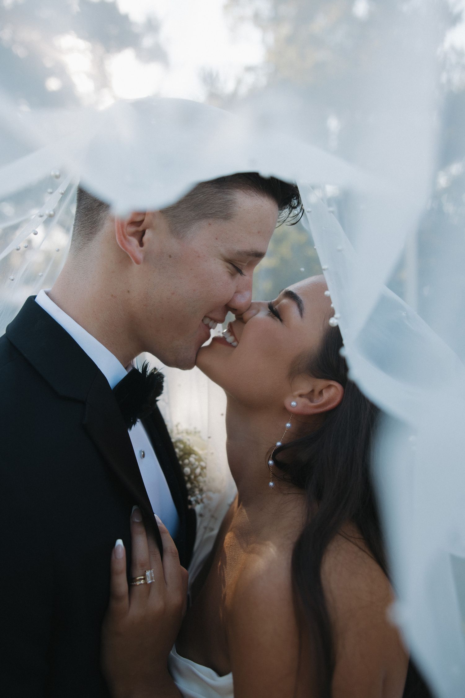 Romantic bride and groom almost kissing under flowing white veil at outdoor wedding celebration.