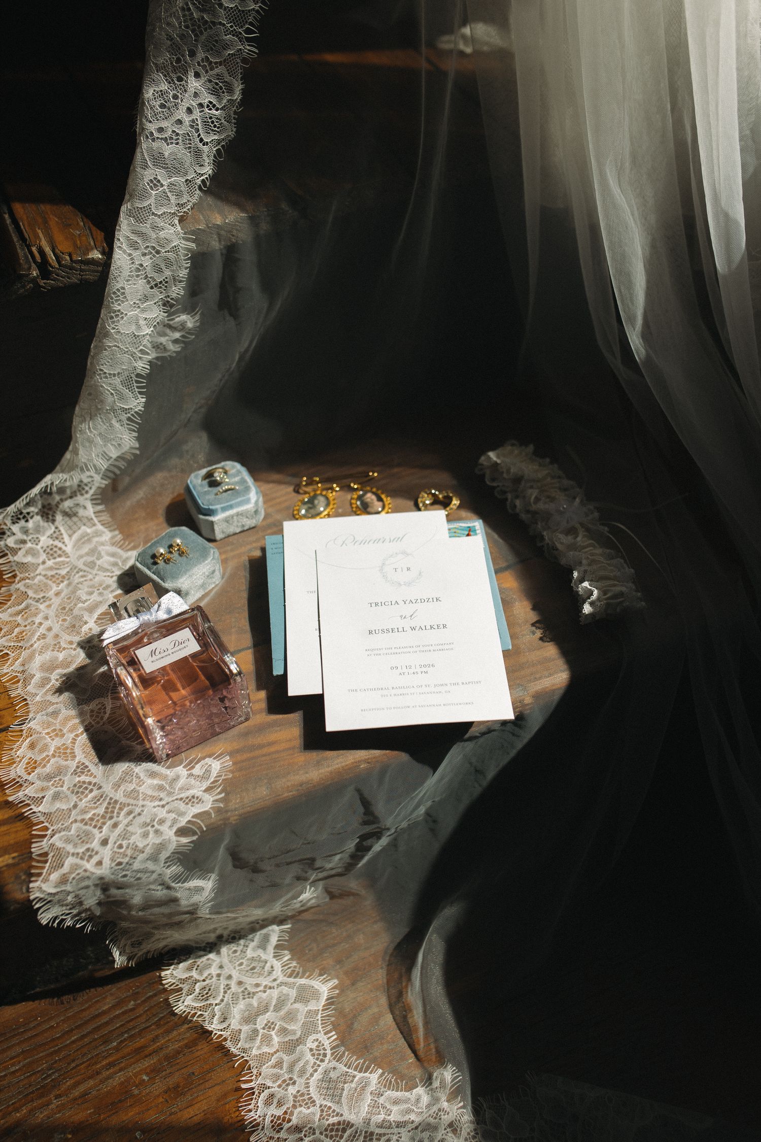 Wedding flat lay with invitation suite, perfume bottle, velvet ring boxes, and lace veil on wooden surface in soft light.