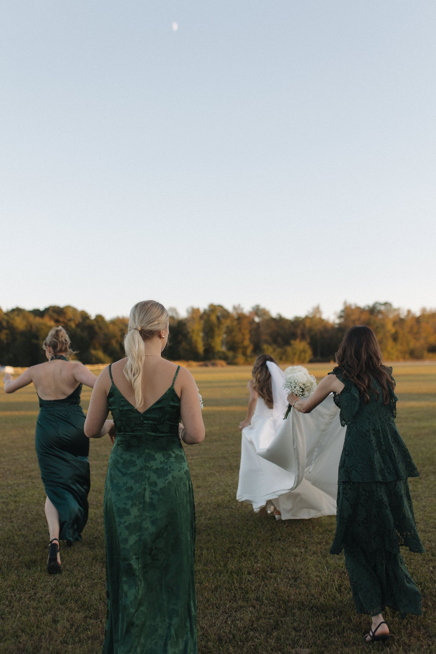 Bridesmaids in green velvet gowns helping bride with white dress as they walk across a sunlit field at golden hour.