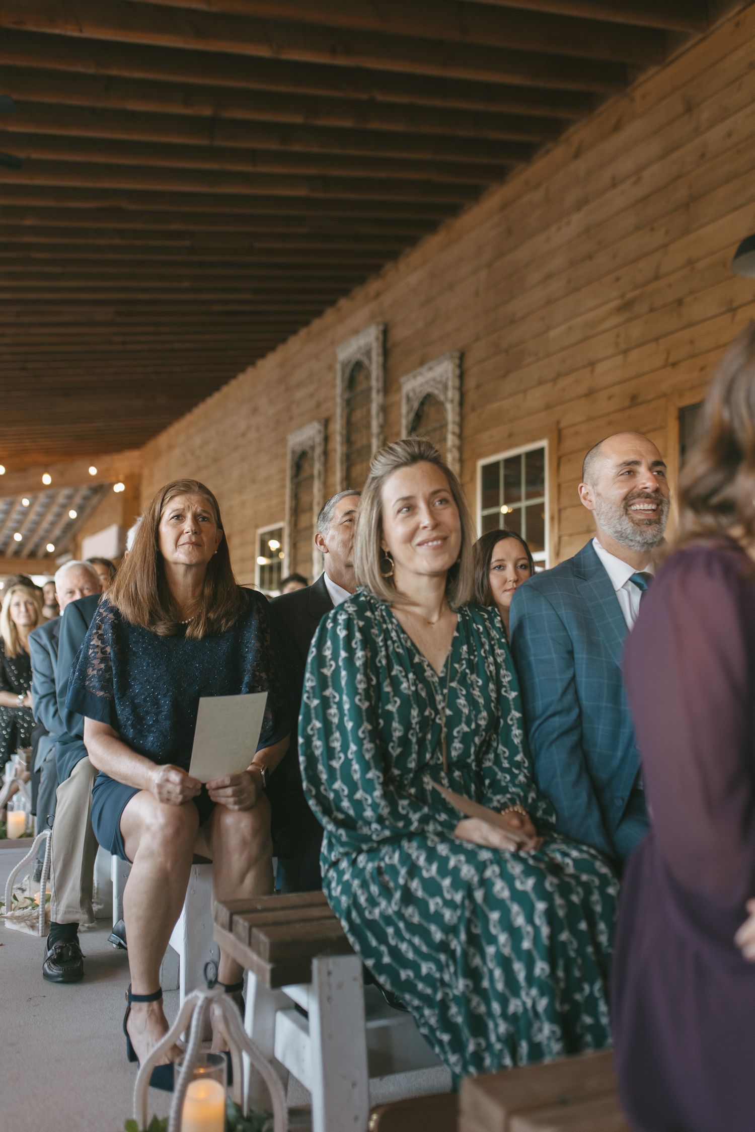 Wedding guests seated on benches in a rustic barn venue, smiling during an indoor ceremony.