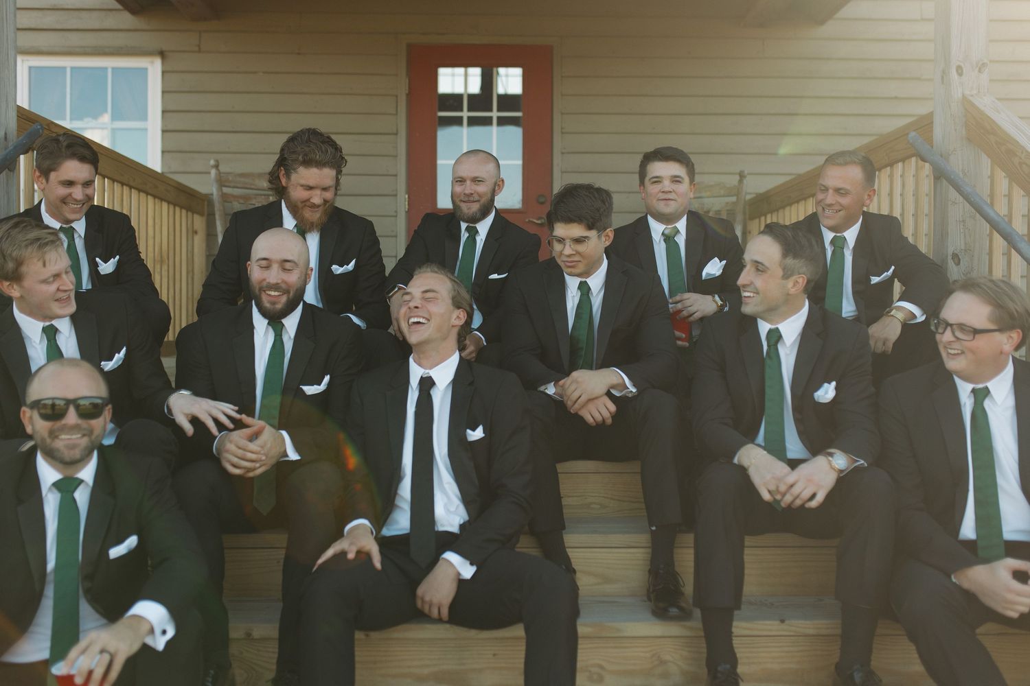 Groomsmen in black suits with green ties laughing on outdoor steps at a wedding venue.