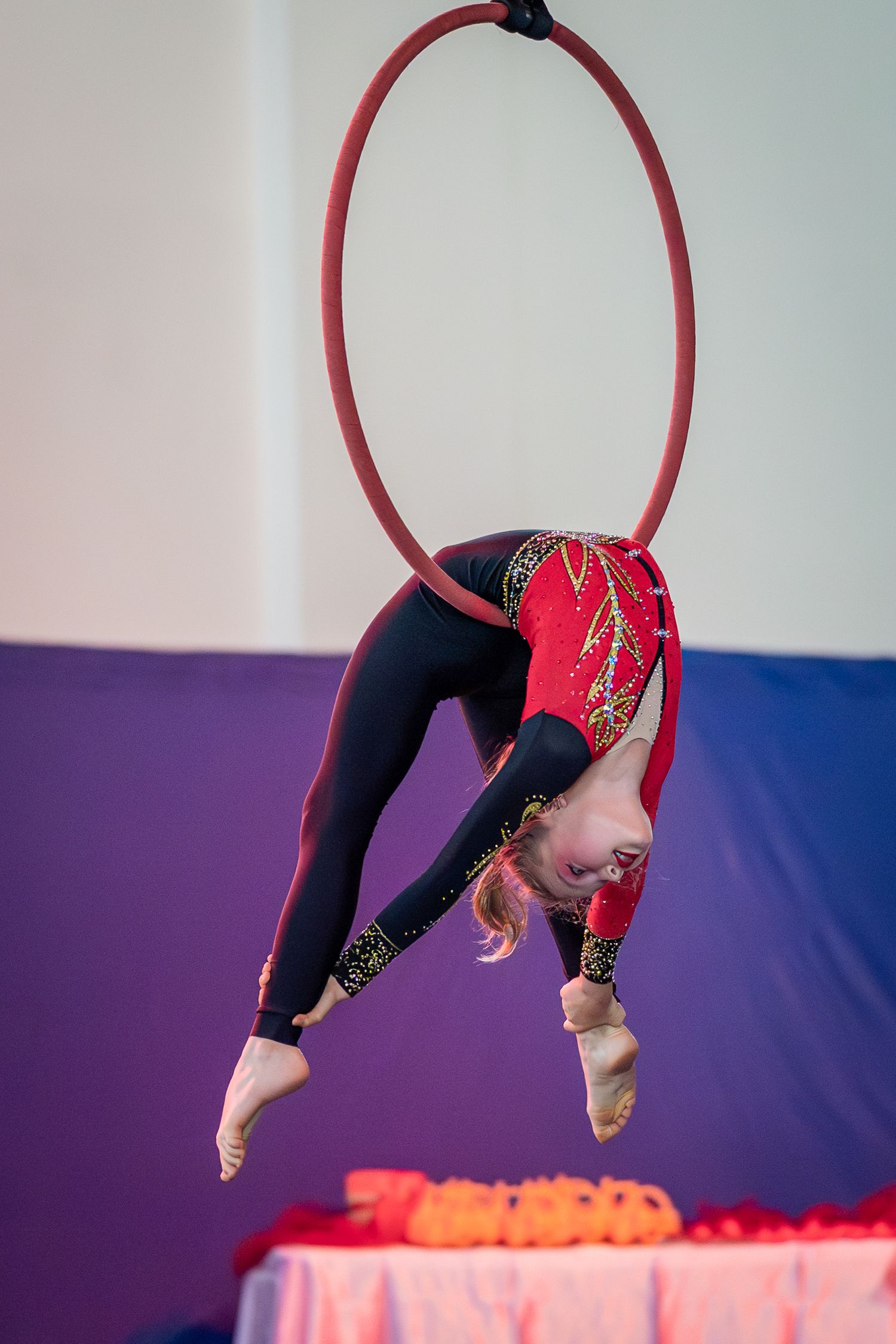 Young girl in red leotard performing aerial hoop acrobatics at gymnastics competition.