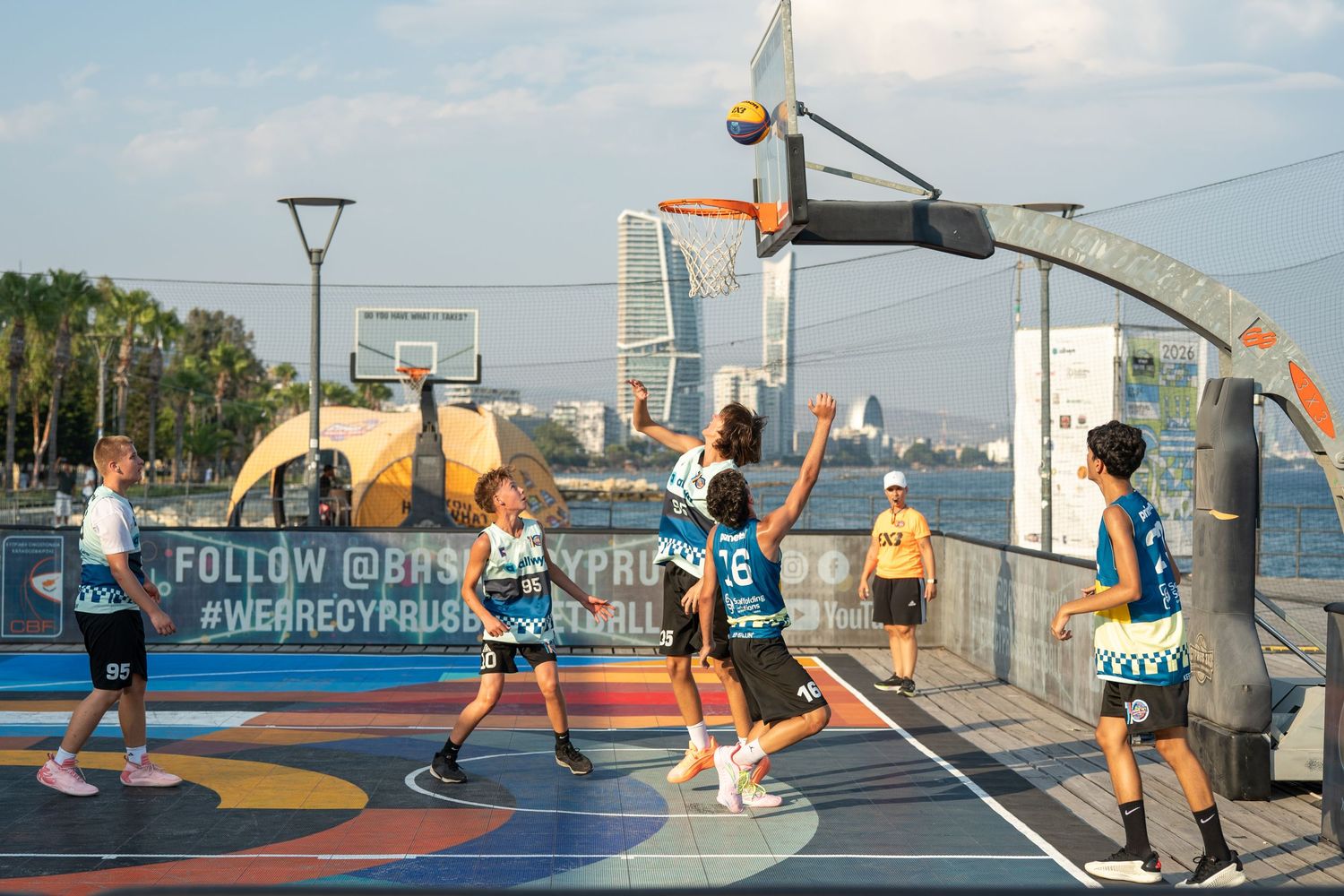 Players competing in 3x3 outdoor basketball tournament on colorful seaside court at sunset.