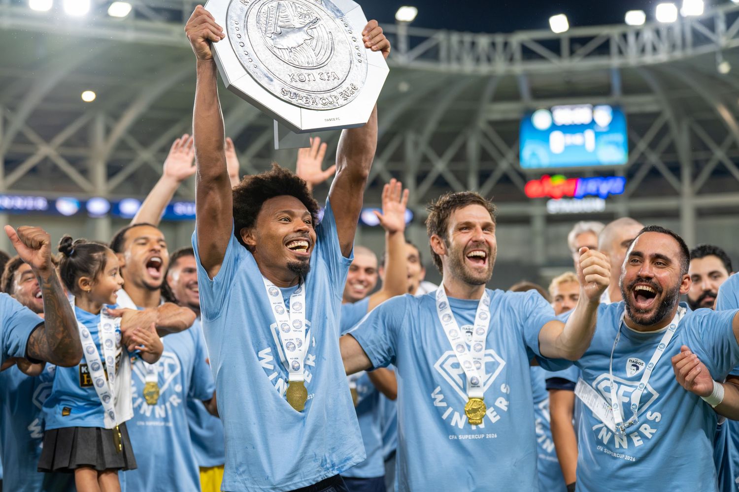 Soccer players in light blue jerseys celebrating a championship trophy lift on the field.