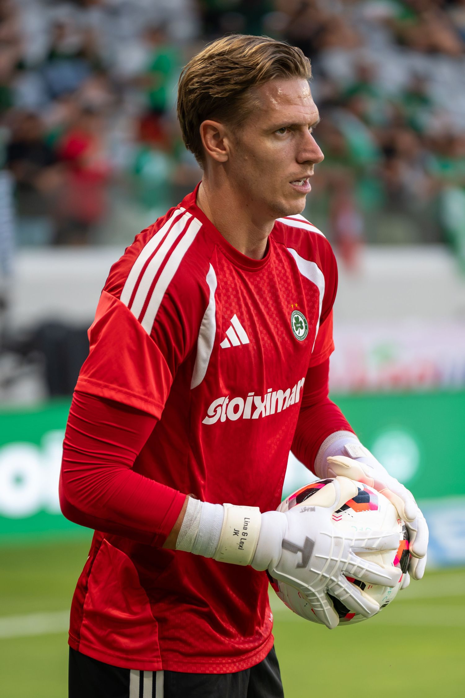 Goalkeeper in red Adidas kit holding a match ball during a professional football game.