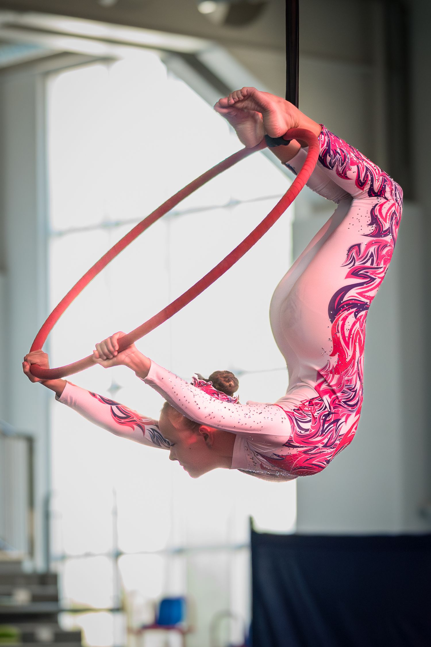 Young gymnast in pink leotard performs aerial hoop routine, bending backwards gracefully during competition.