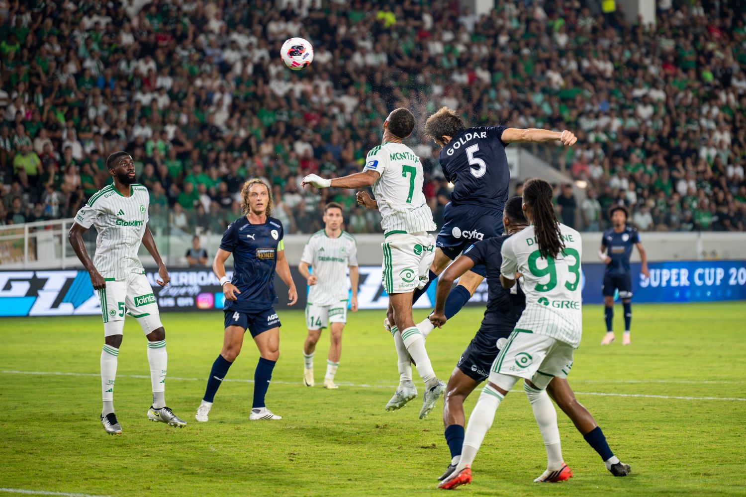 Soccer players competing for header during a Super Cup match in a green and white stadium.
