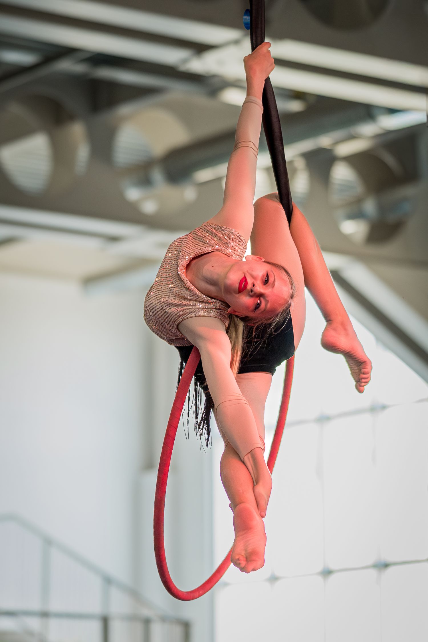 Young female aerialist in sequined leotard performing splits on aerial hoop indoors.
