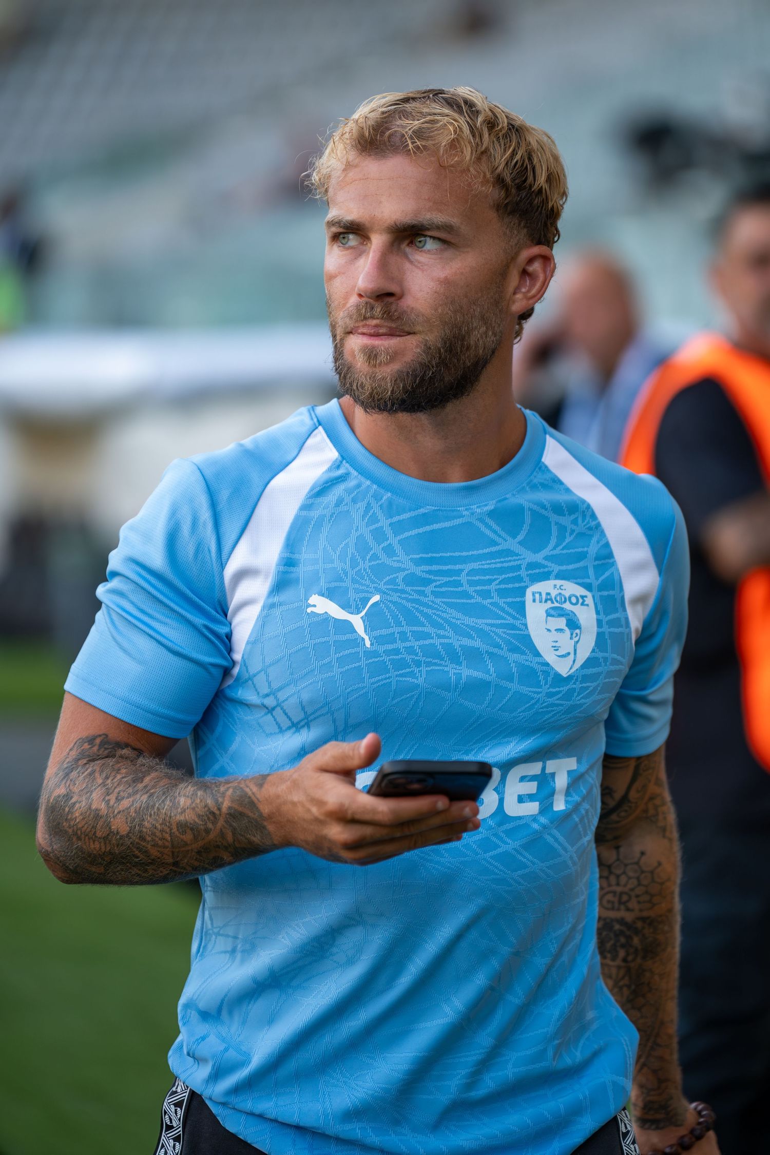 Tattooed blonde footballer in light blue Pafos FC kit holding a phone at the stadium.
