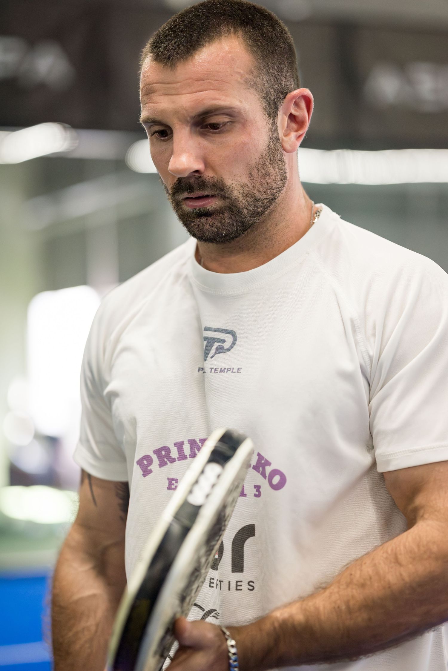 Athletic man in white Primo Tempo t-shirt holding a padel racket indoors at sports facility.