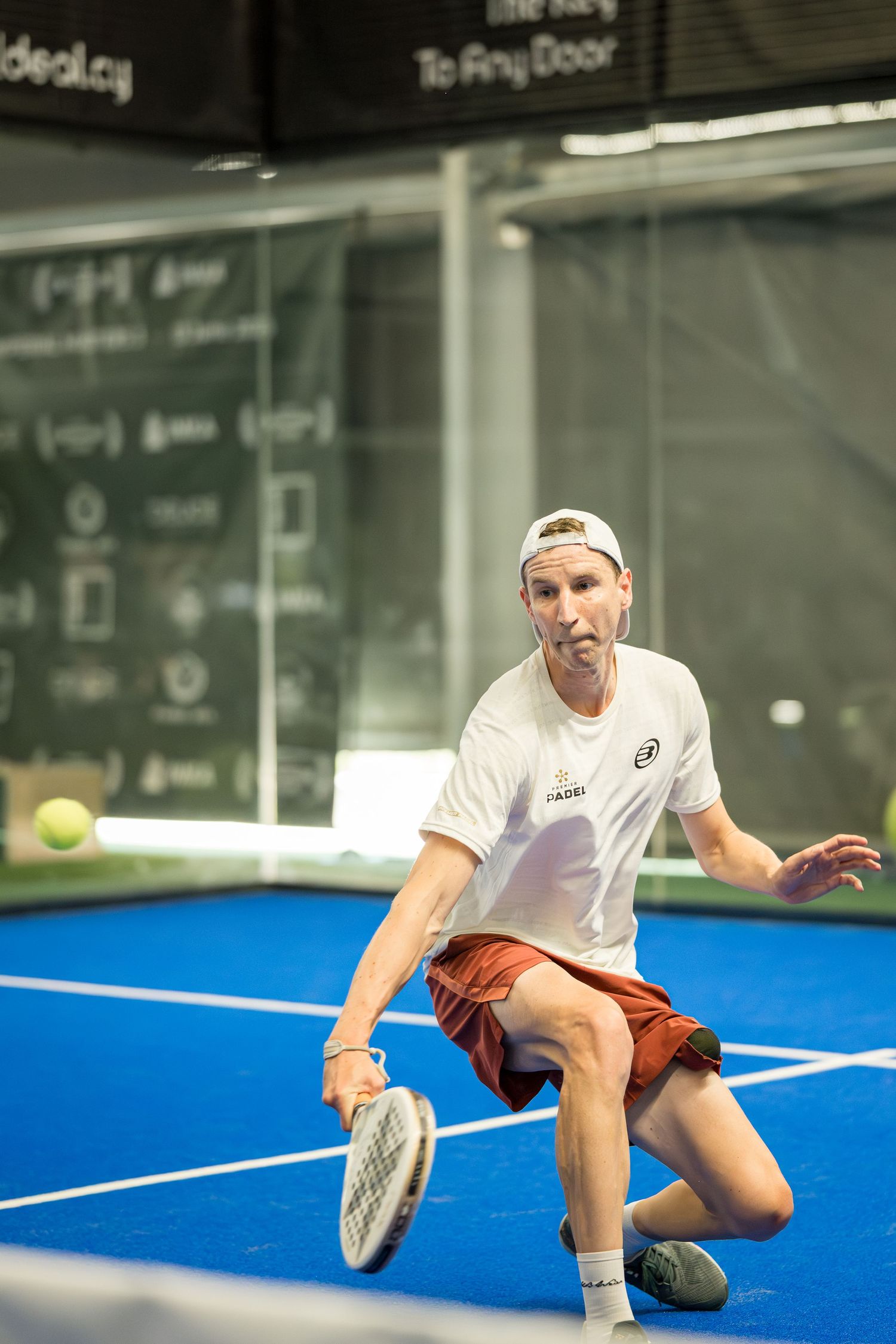 Male padel player in white shirt lunging low to return ball on indoor blue court.
