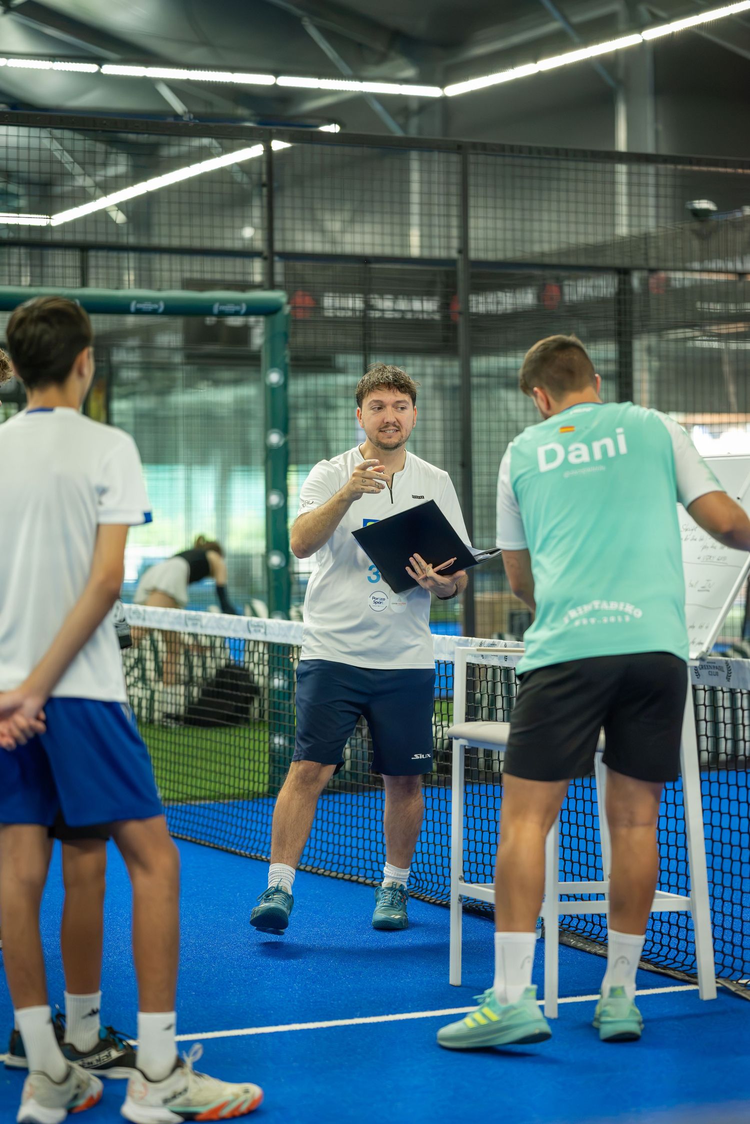 Indoor padel coach holding a clipboard and instructing two players near the net on a blue court.