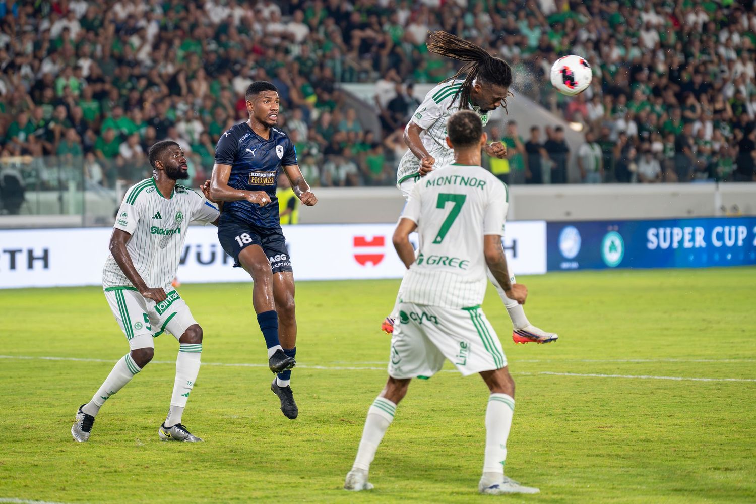 Soccer players compete for a header during a Super Cup match between teams in green and navy blue uniforms.