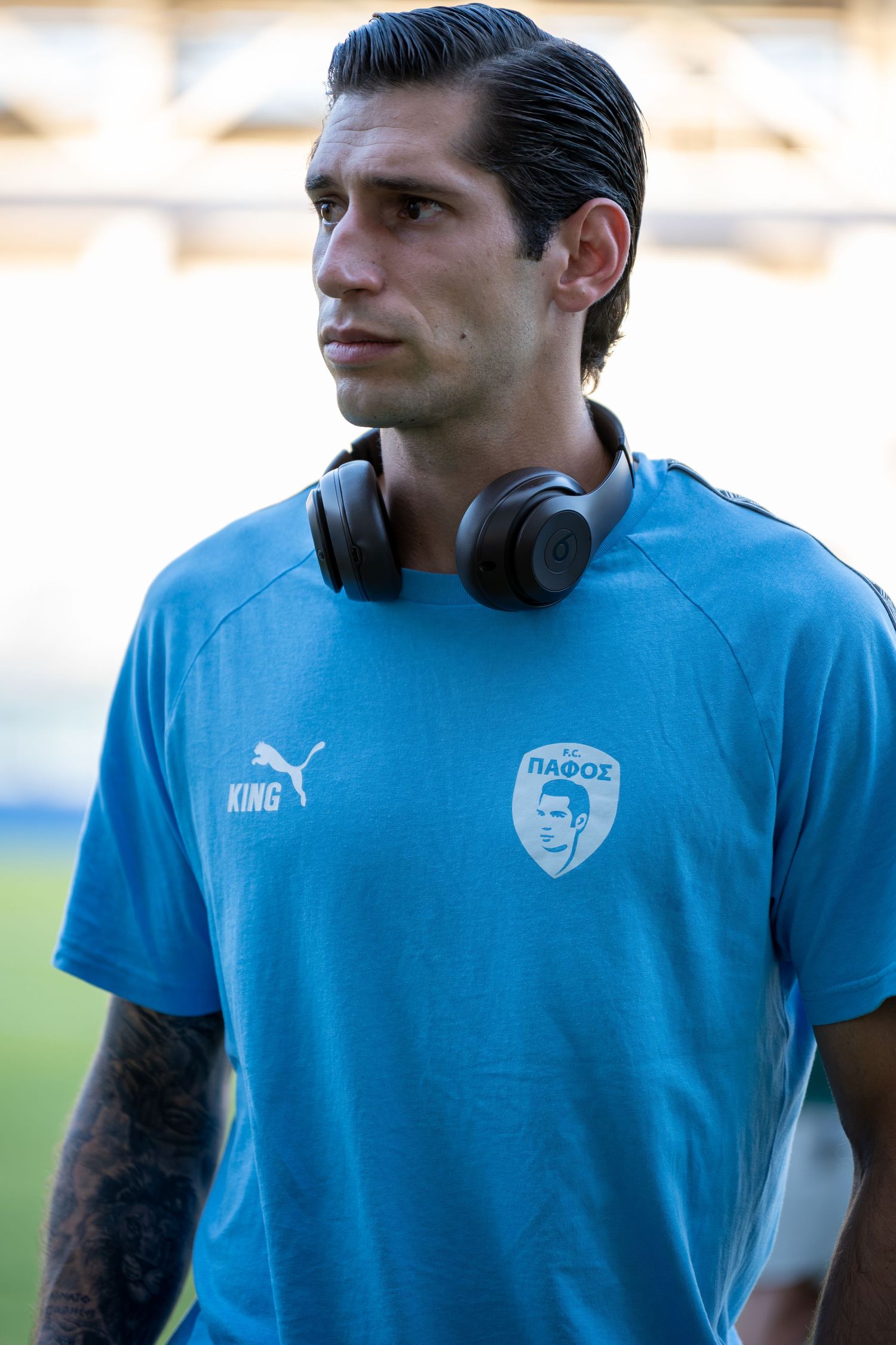 Male footballer in light blue Pafos FC Puma shirt with headphones around neck at stadium.