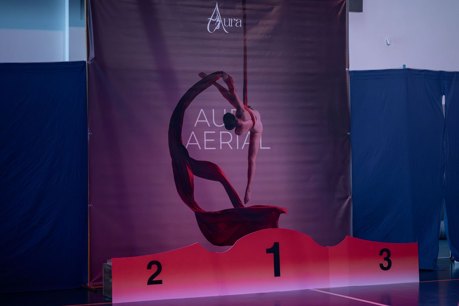 Aerial acrobat performing a graceful pose on silk ribbons above a first-place podium on stage.