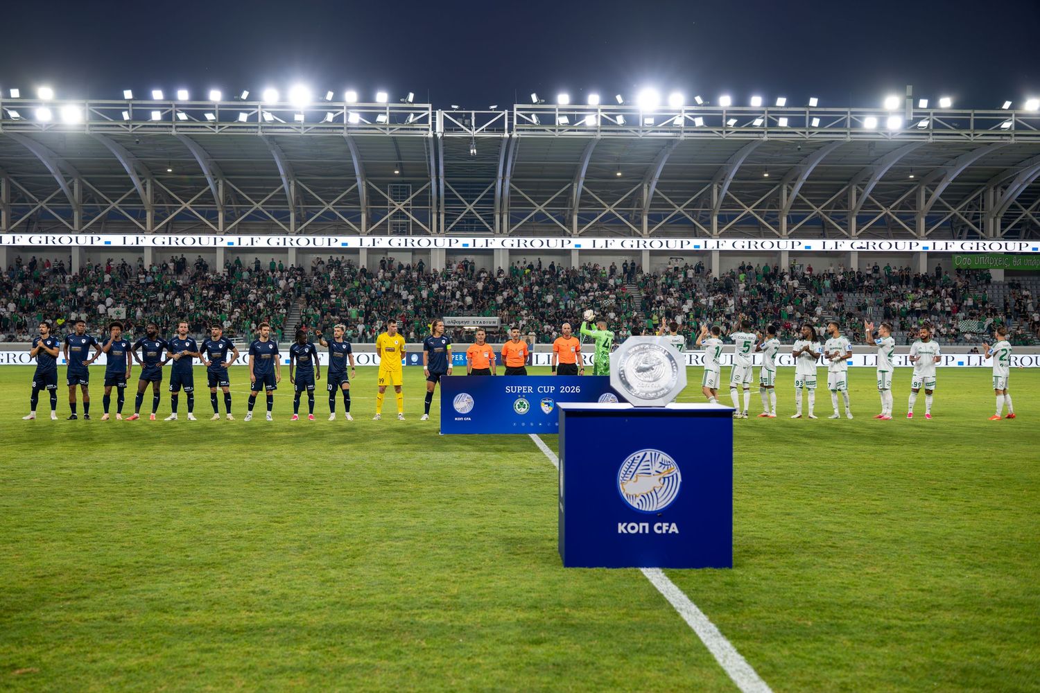 Soccer teams lined up on a floodlit stadium pitch during a KOP CFA cup match at night.