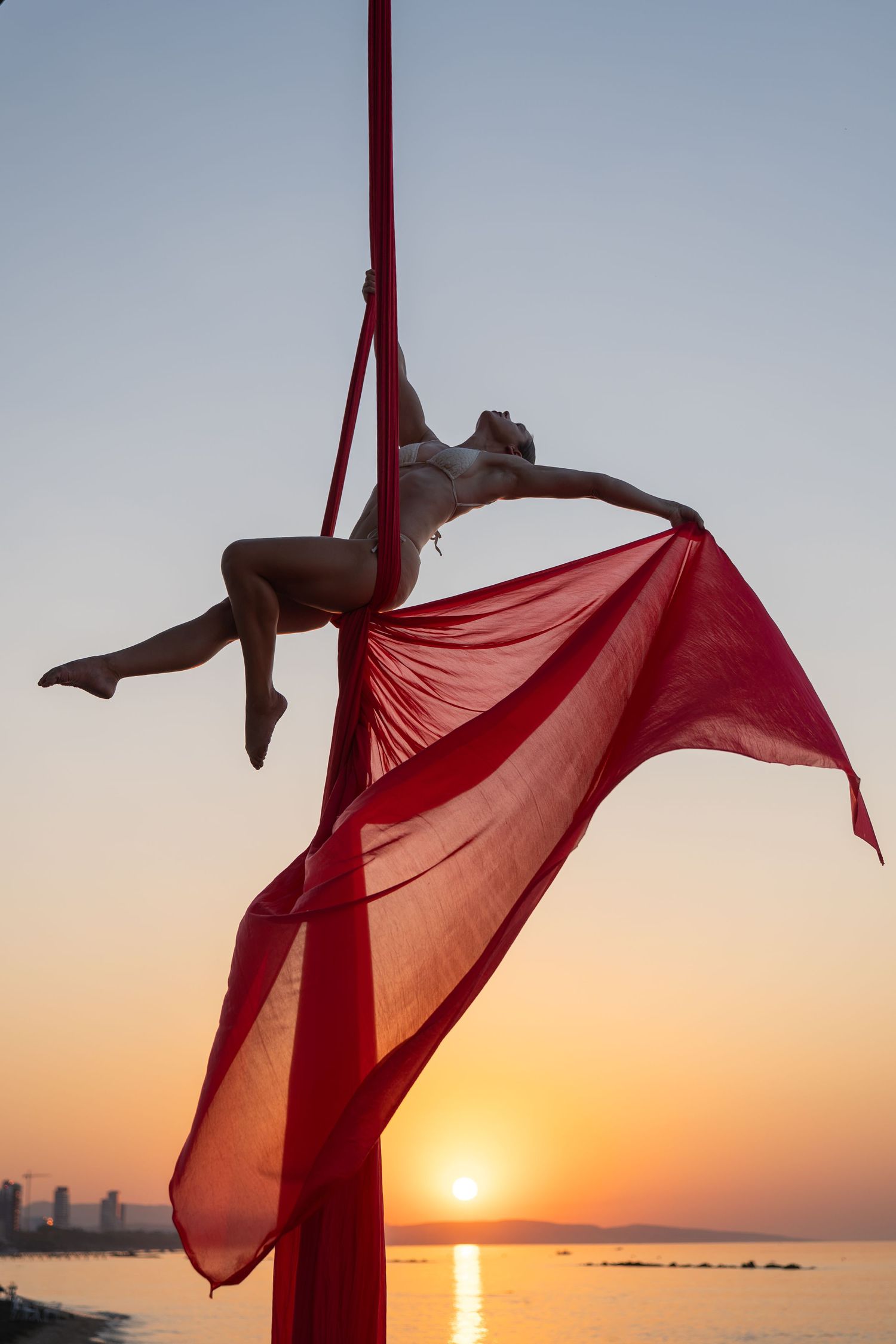 Female aerialist performing graceful pose on red aerial silks at sunset over water.