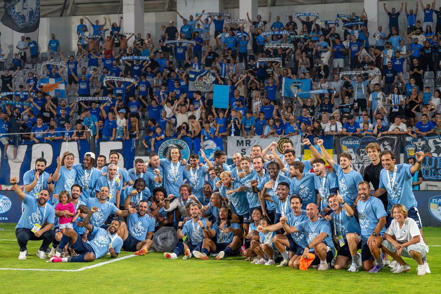Football team and staff posing together for a group photo in front of blue-shirted fans in a stadium.