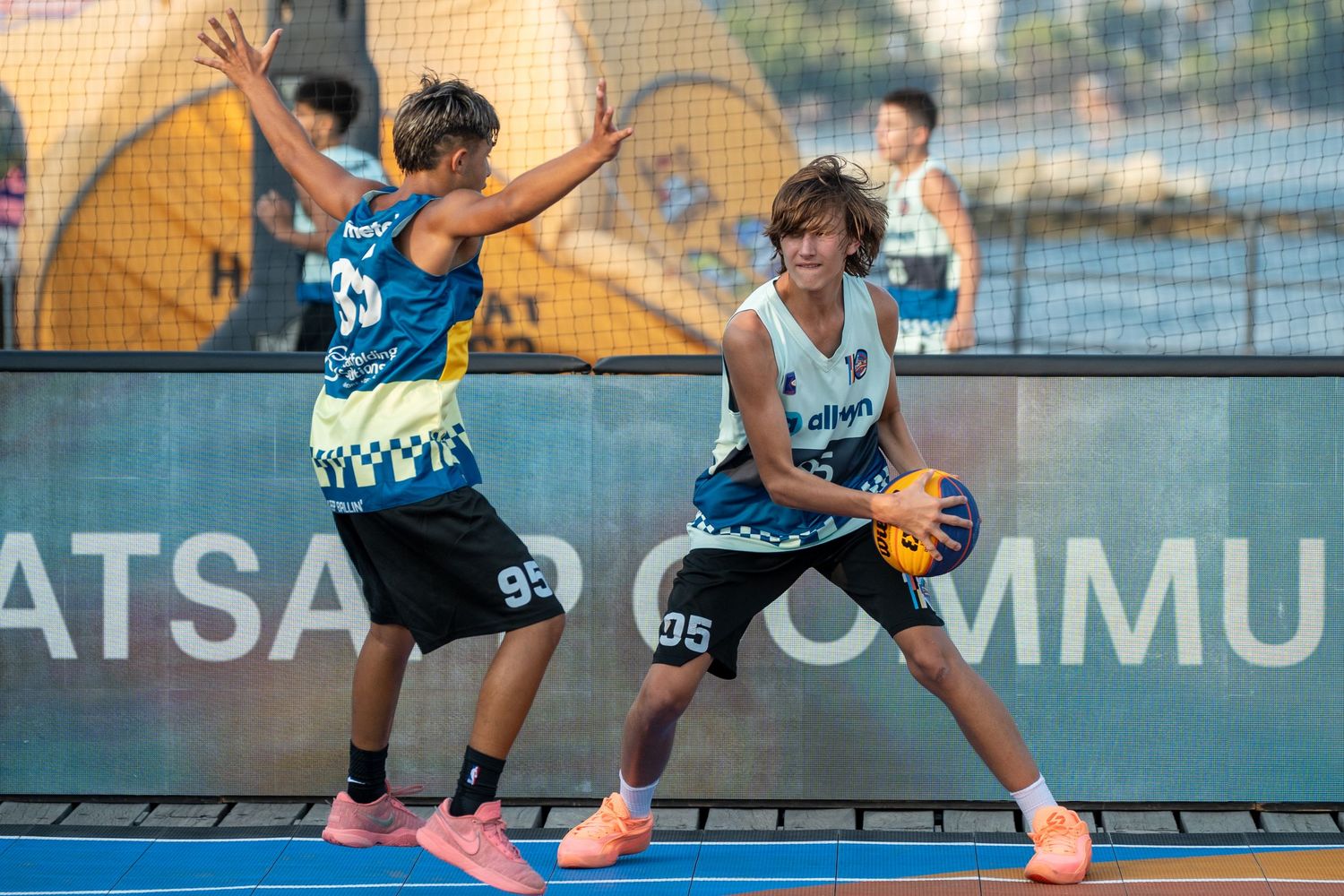 Two young basketball players compete in a 3x3 street basketball game, one dribbling while the other defends.