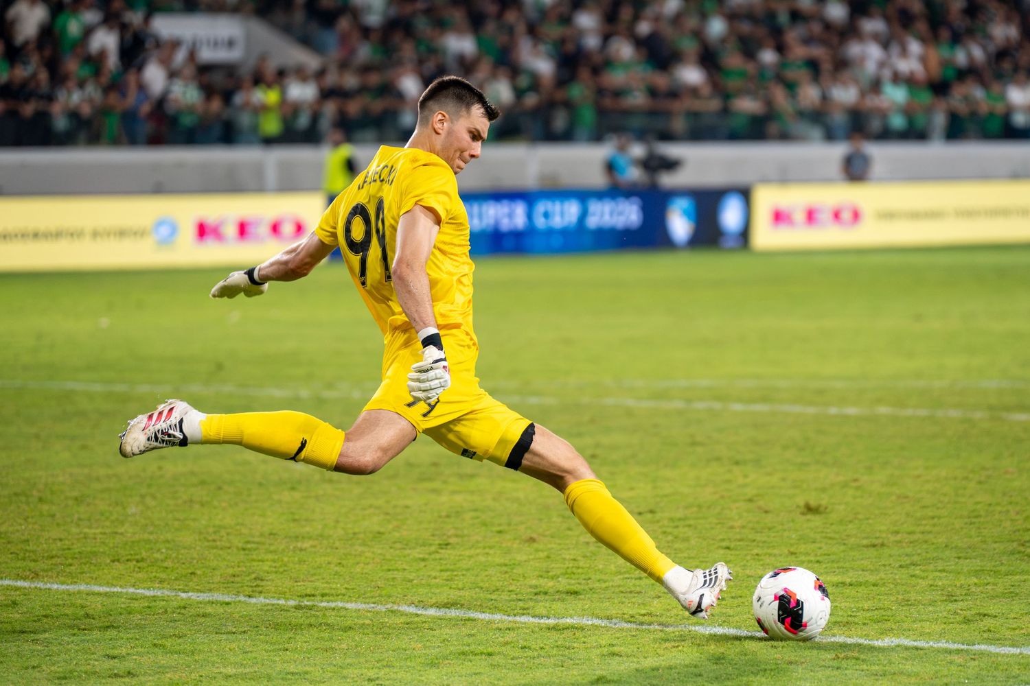 Goalkeeper in yellow kit taking a goal kick during a Super Cup football match at a stadium.