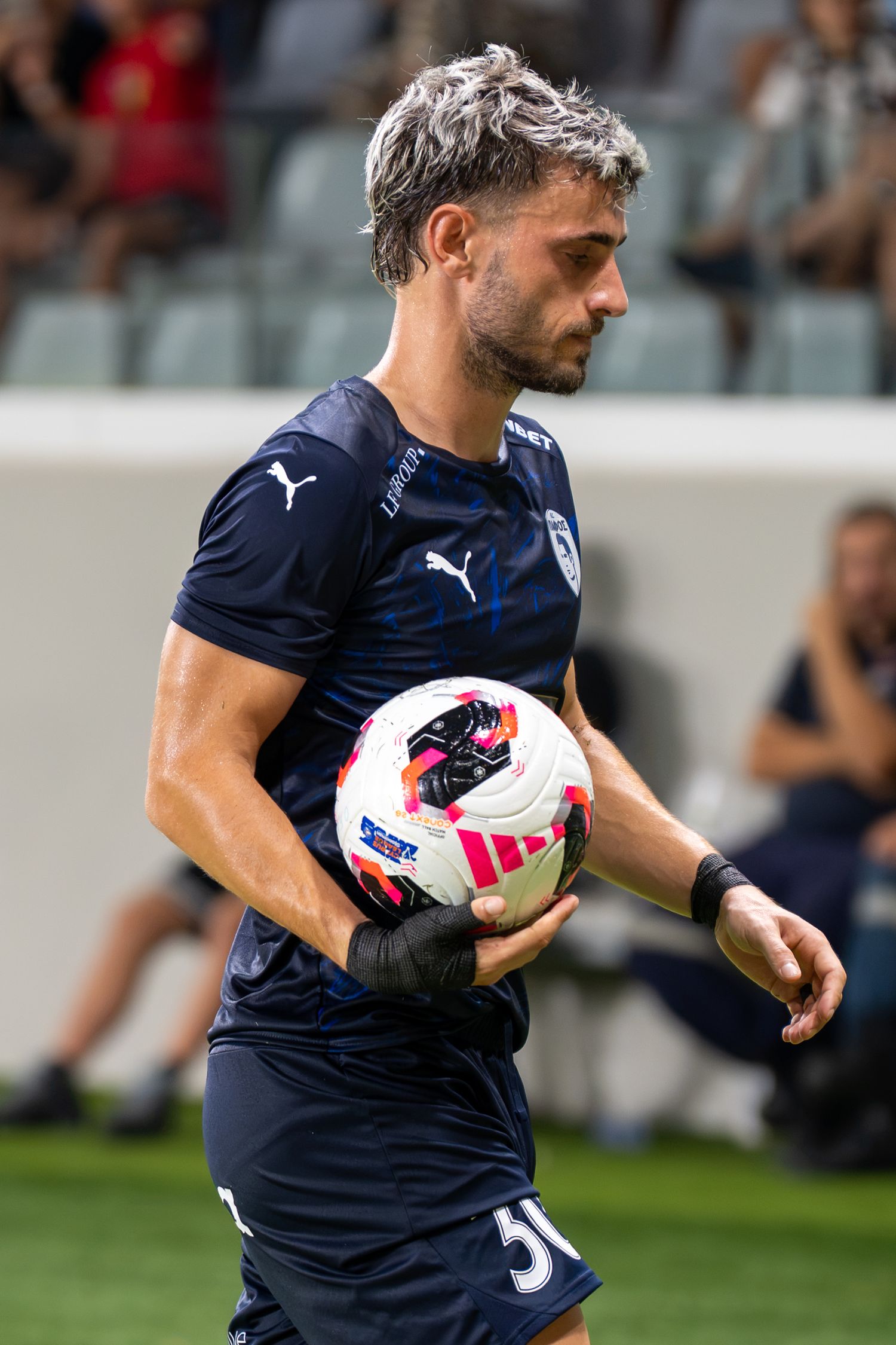 Male soccer player in dark navy Puma jersey holding match ball on illuminated football pitch.