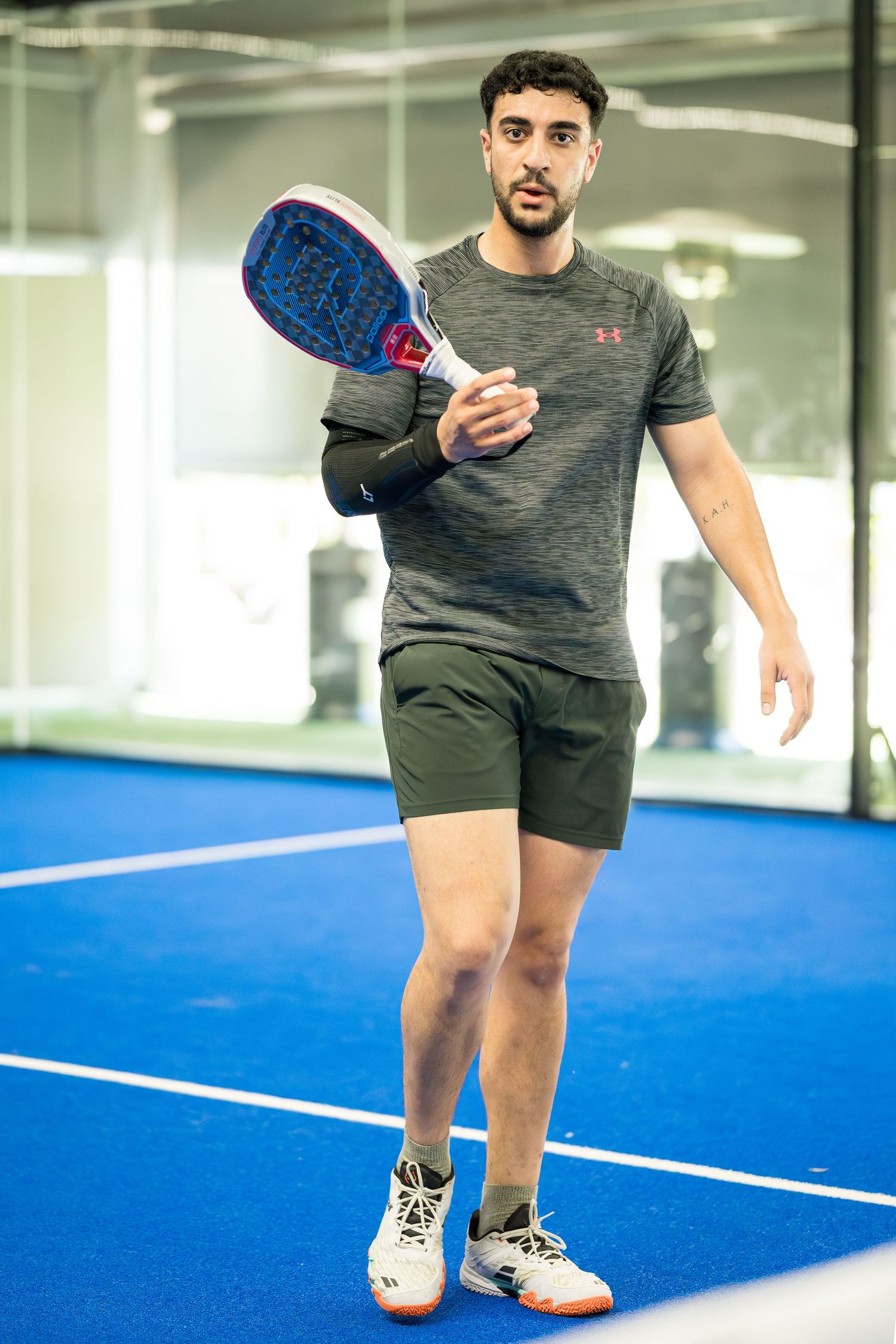 Young man in grey Under Armour shirt holding a padel racket on a blue indoor court.