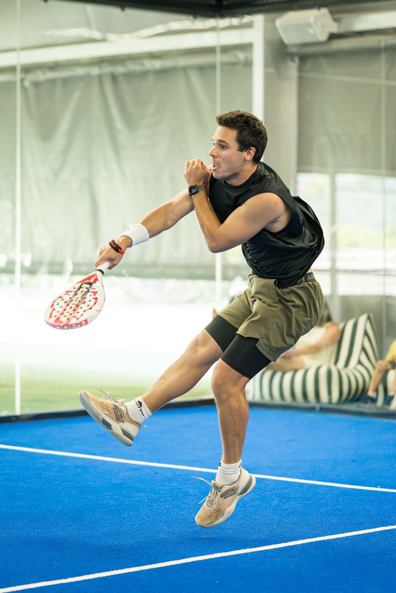 Athletic man in black vest jumping mid-air to hit ball with padel racket on blue court.