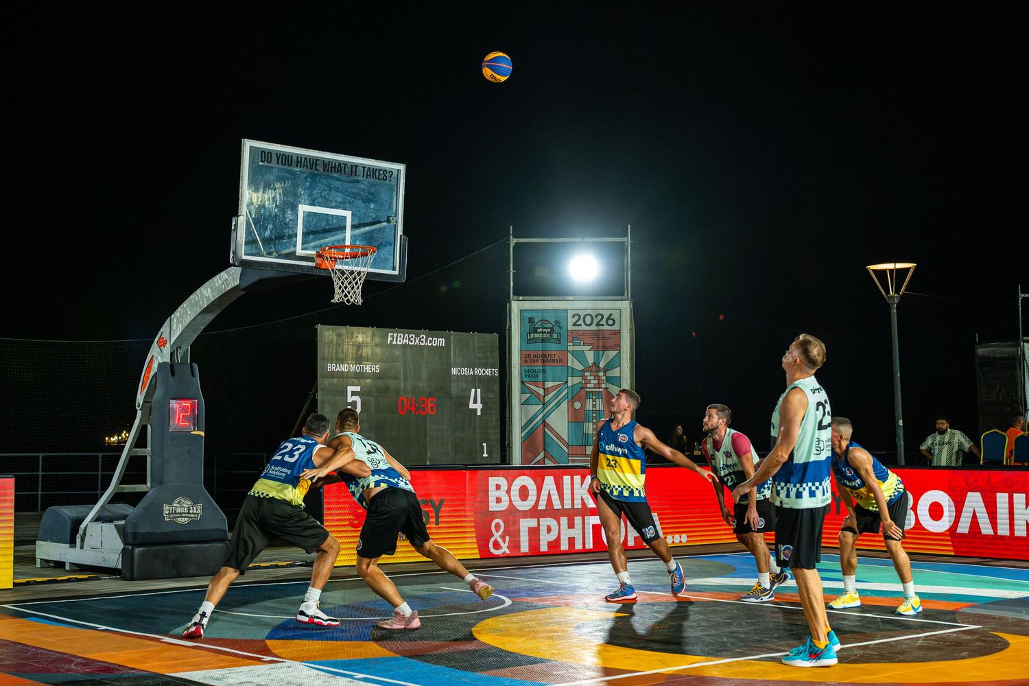 Athletes compete in 3x3 basketball tournament under floodlights on outdoor court at night.
