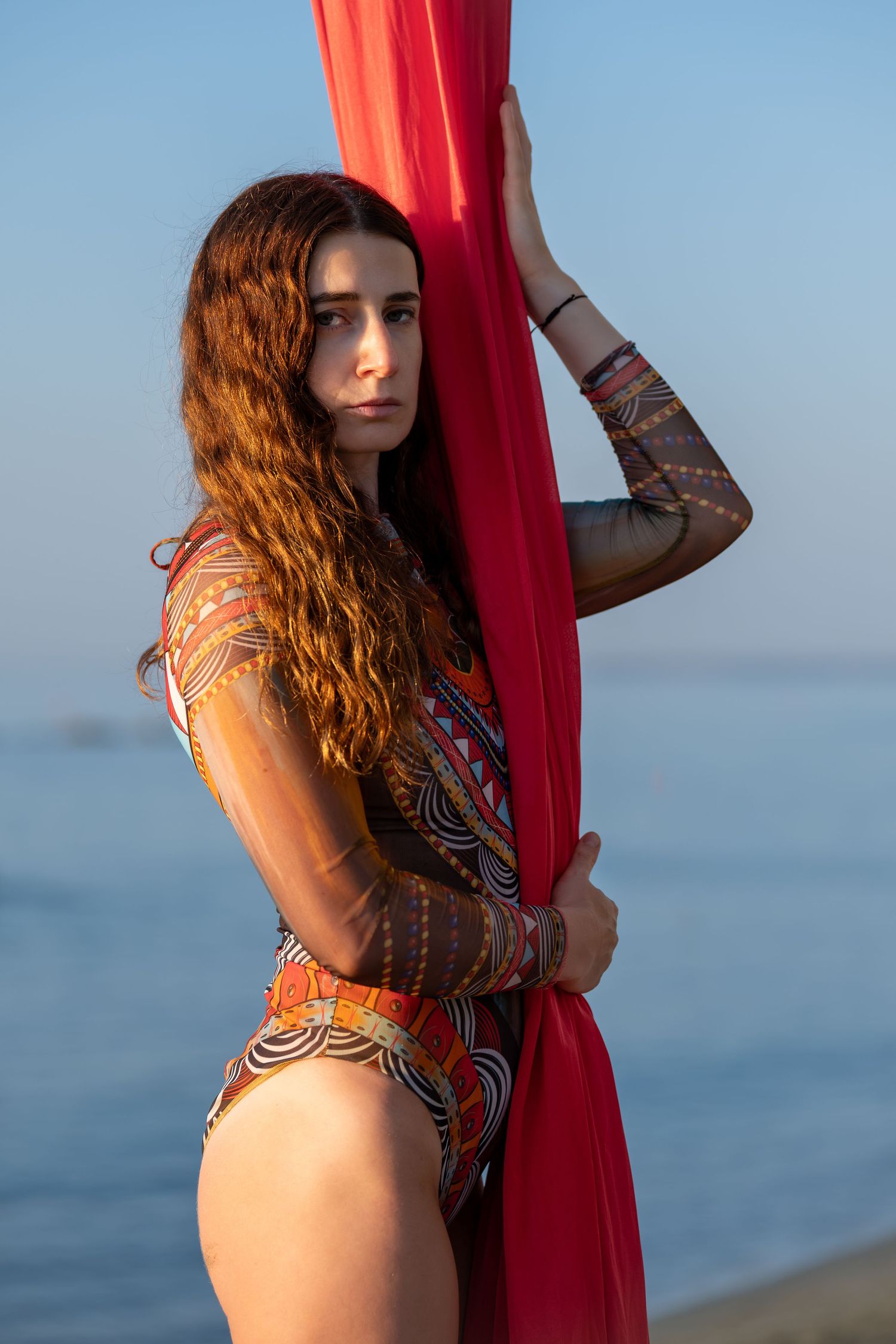 Young woman in colorful patterned swimsuit holding red aerial silk fabric on beach.