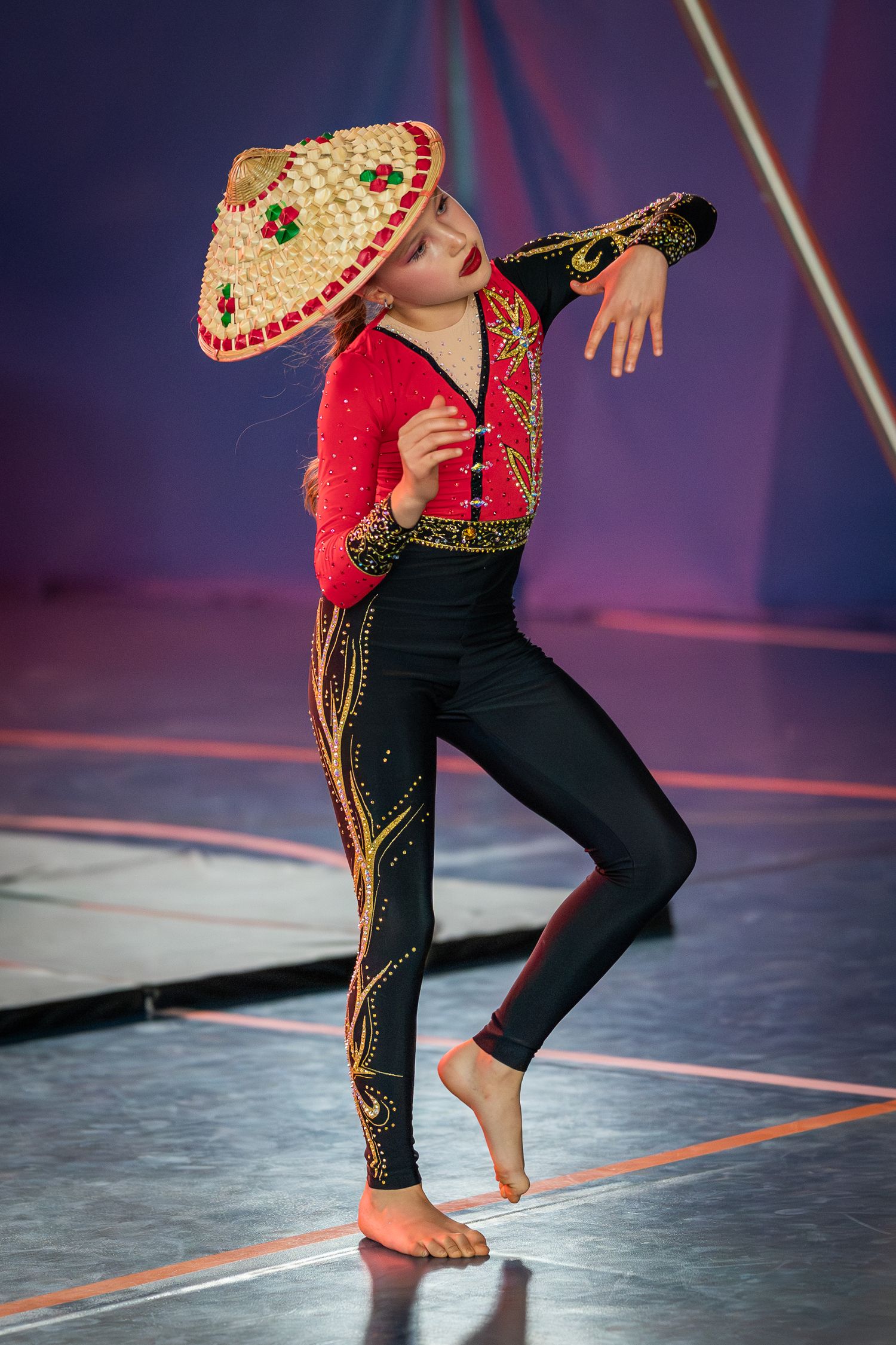 Young girl in red costume and sombrero performing dance routine on stage barefoot.