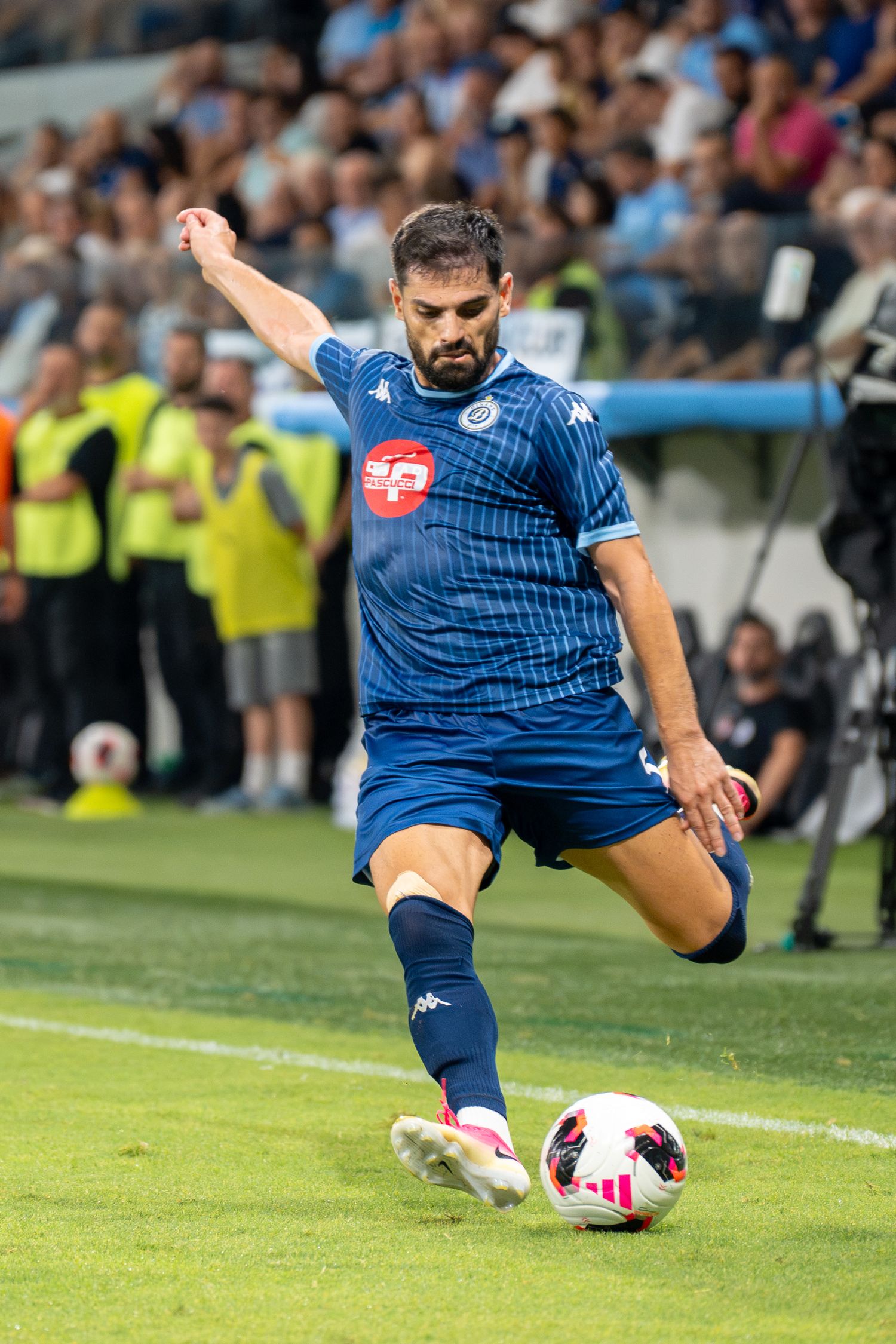 Soccer player in blue Kappa kit strikes the ball during a match with stadium crowd in background.