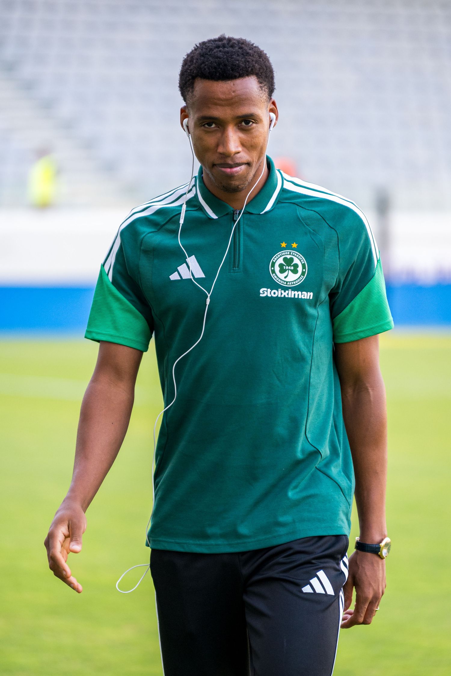 Young footballer in green Adidas Maccabi kit walking on pitch with earphones before a match.