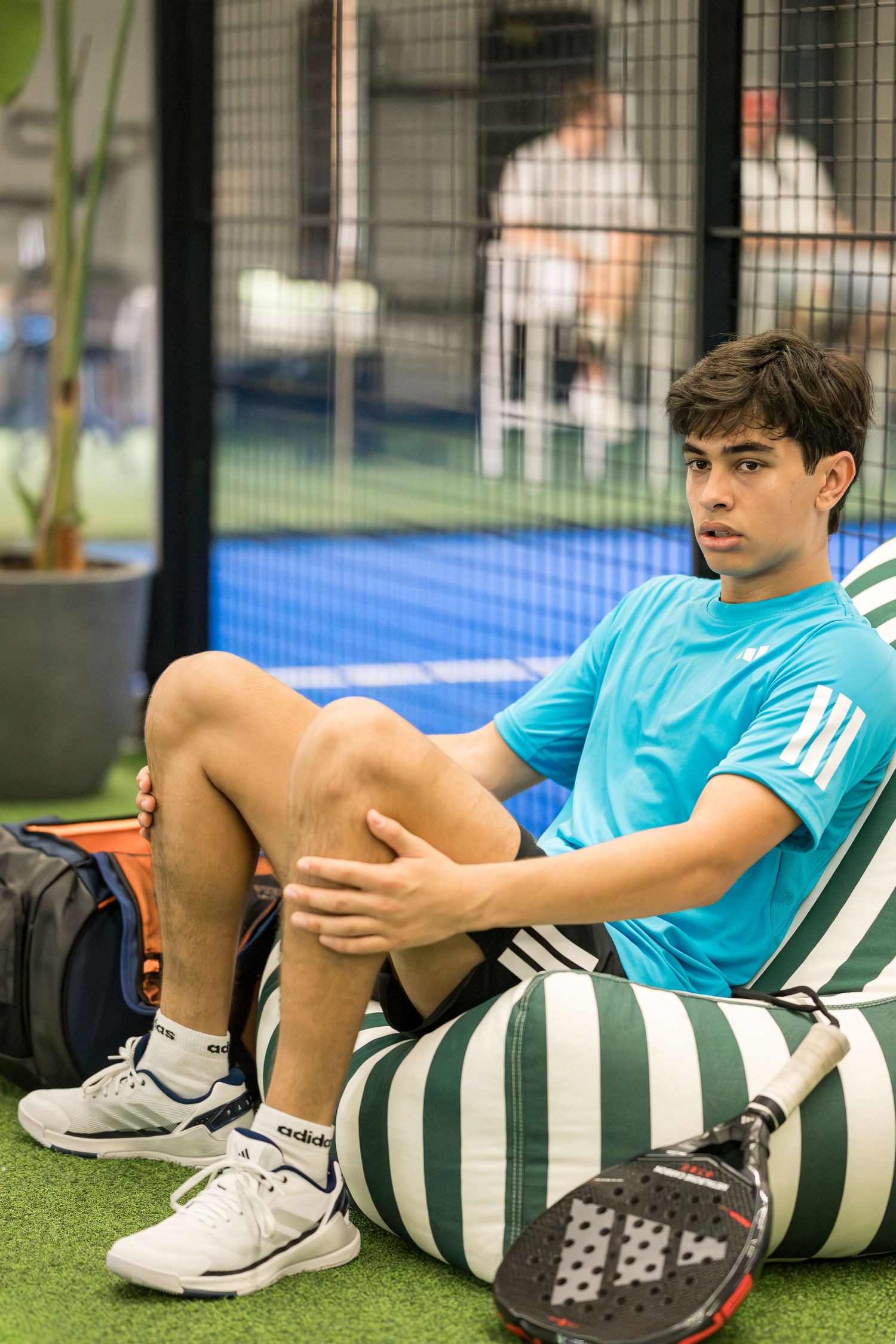 Young male padel player in blue Adidas shirt resting on a bean bag near the court.
