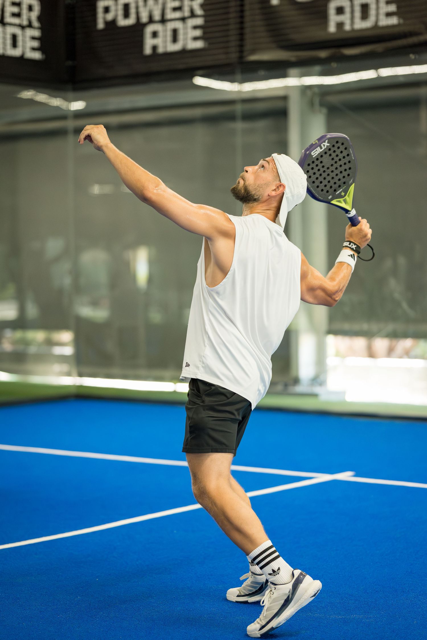 Male padel player in white shirt serving ball on indoor blue court with Powerade signage.