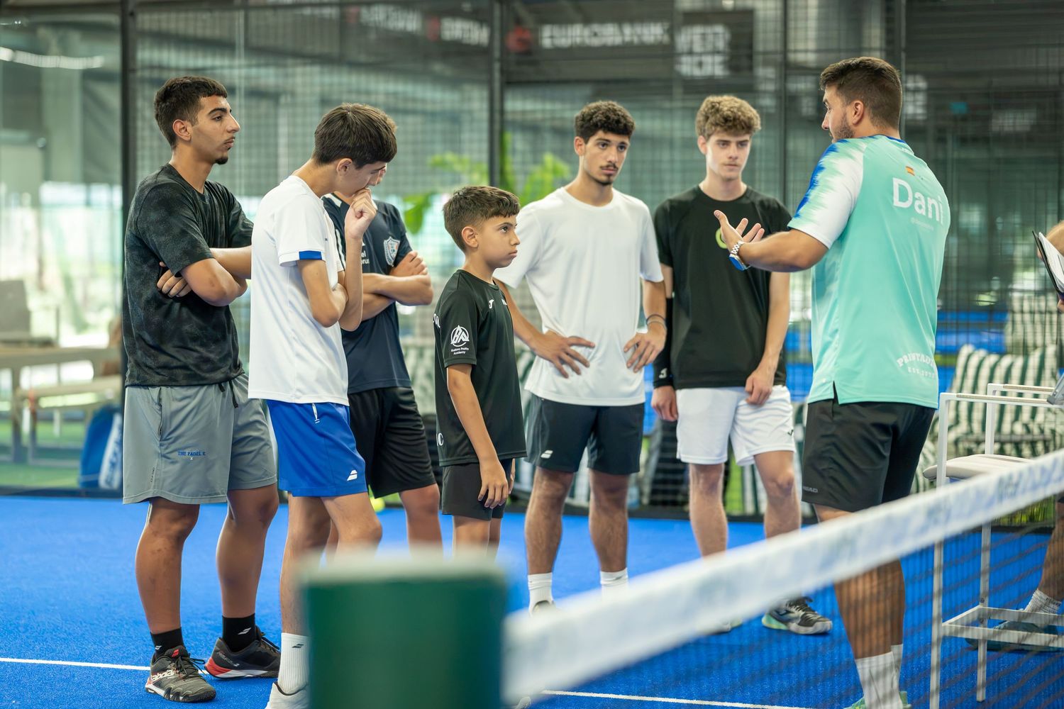 A padel coach instructing a group of young players beside the net on an outdoor court.