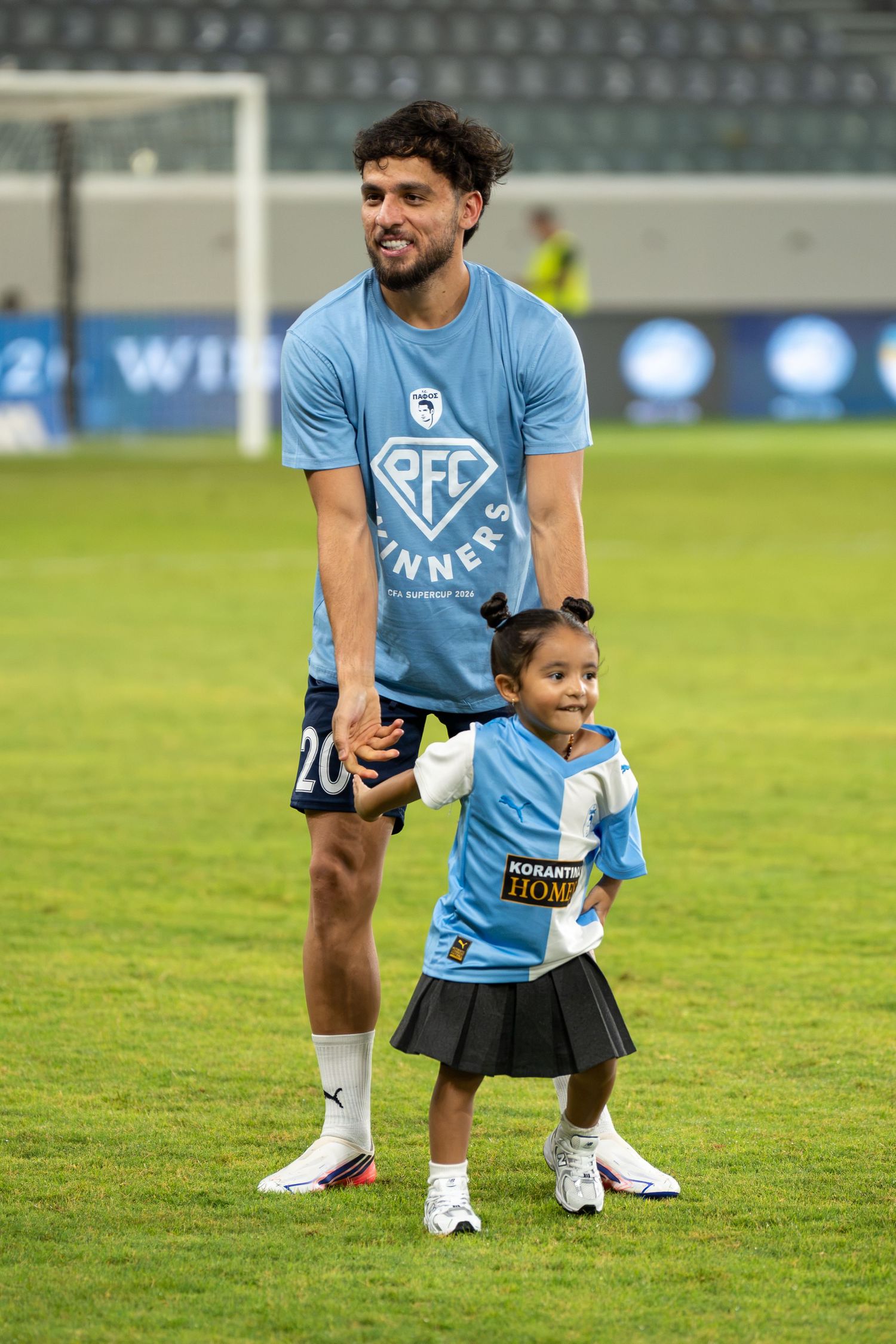 Football player in light blue RFC Winners shirt posing with a young child on the pitch.