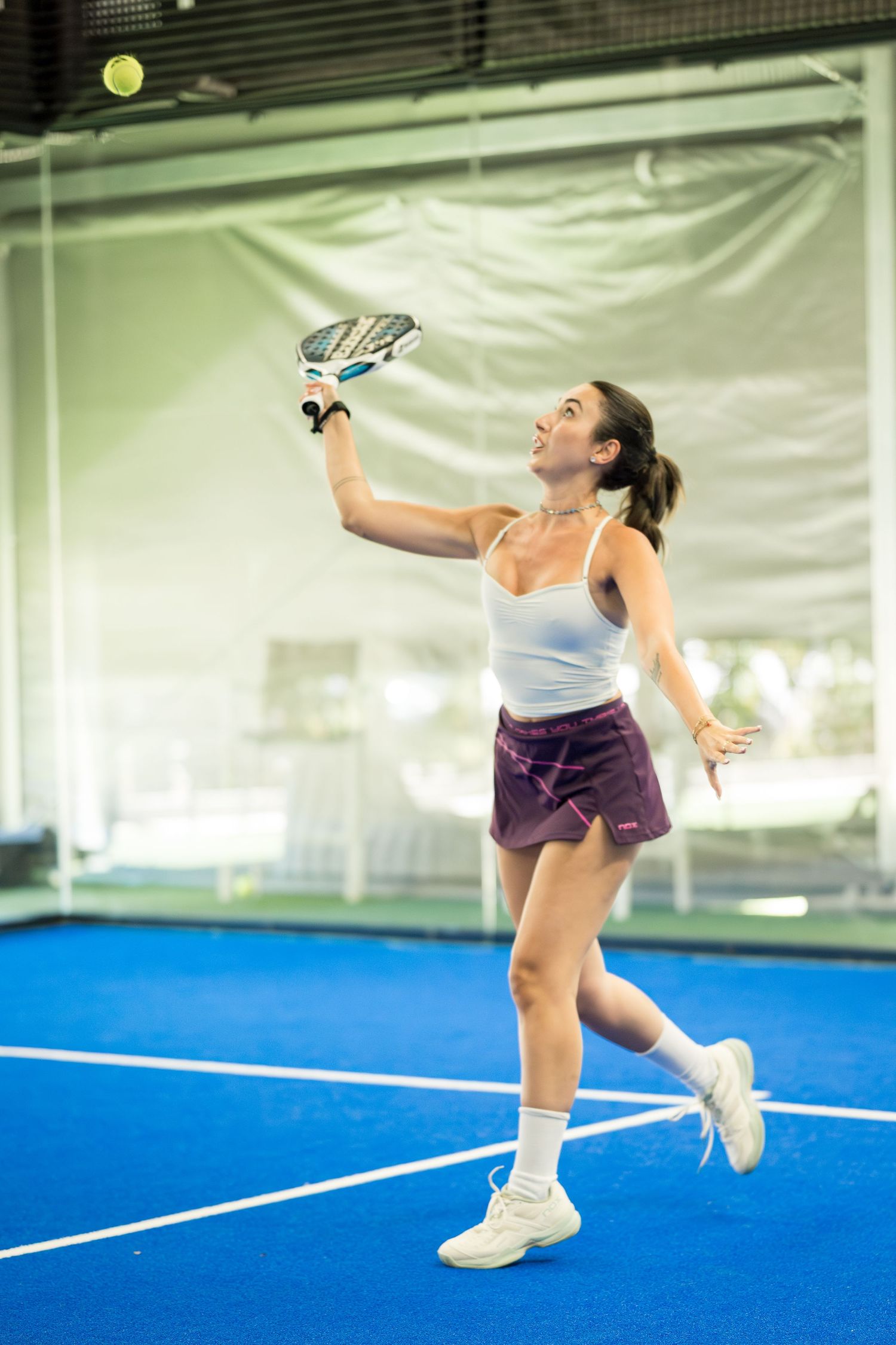 Young woman in white top leaping to smash ball with padel racket on indoor blue court.