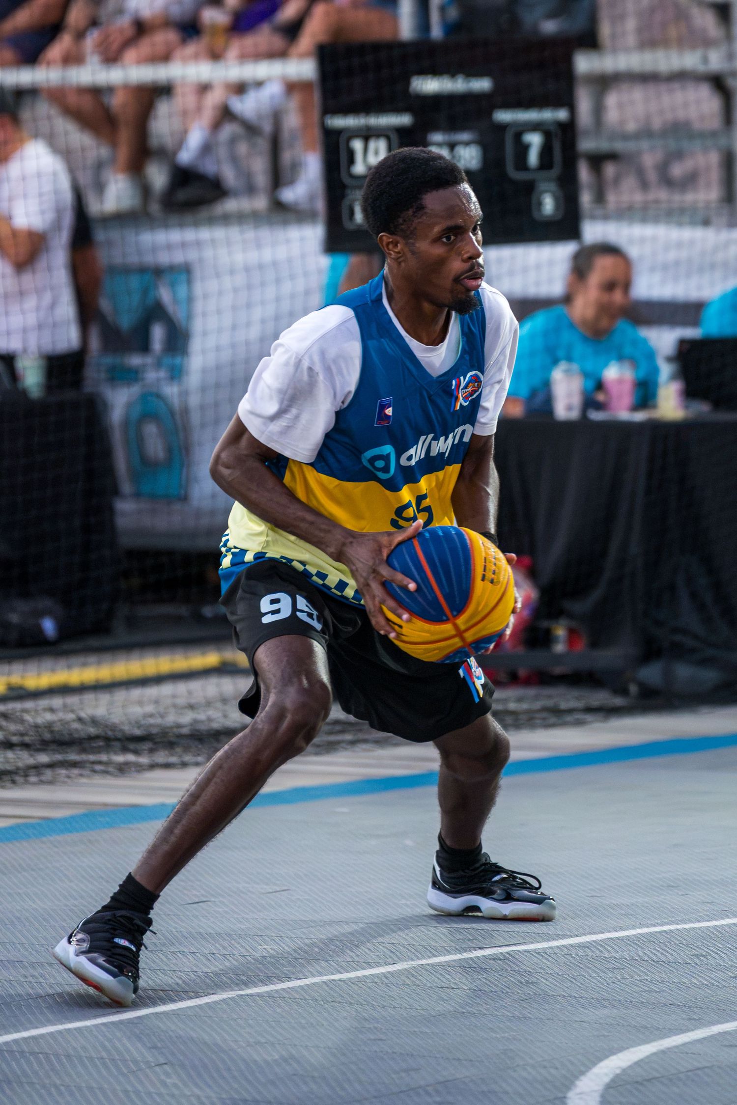 A basketball player in a yellow and blue jersey dribbles the ball during an outdoor 3x3 street basketball event.