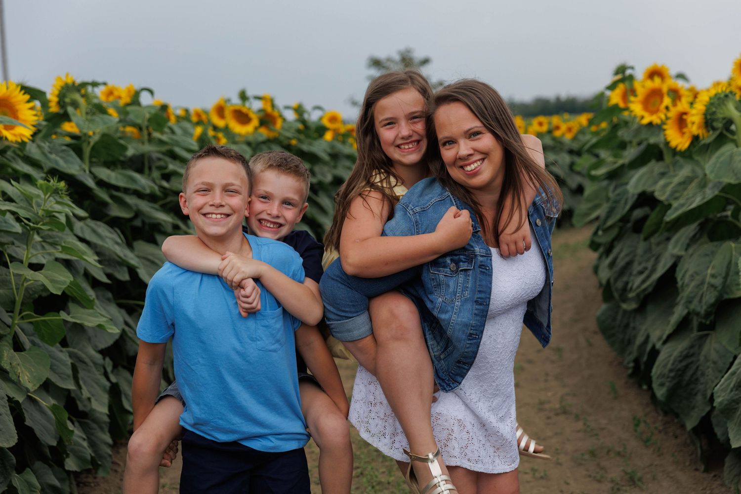 Mother piggyback carrying daughter while son carries brother in sunflower field path.