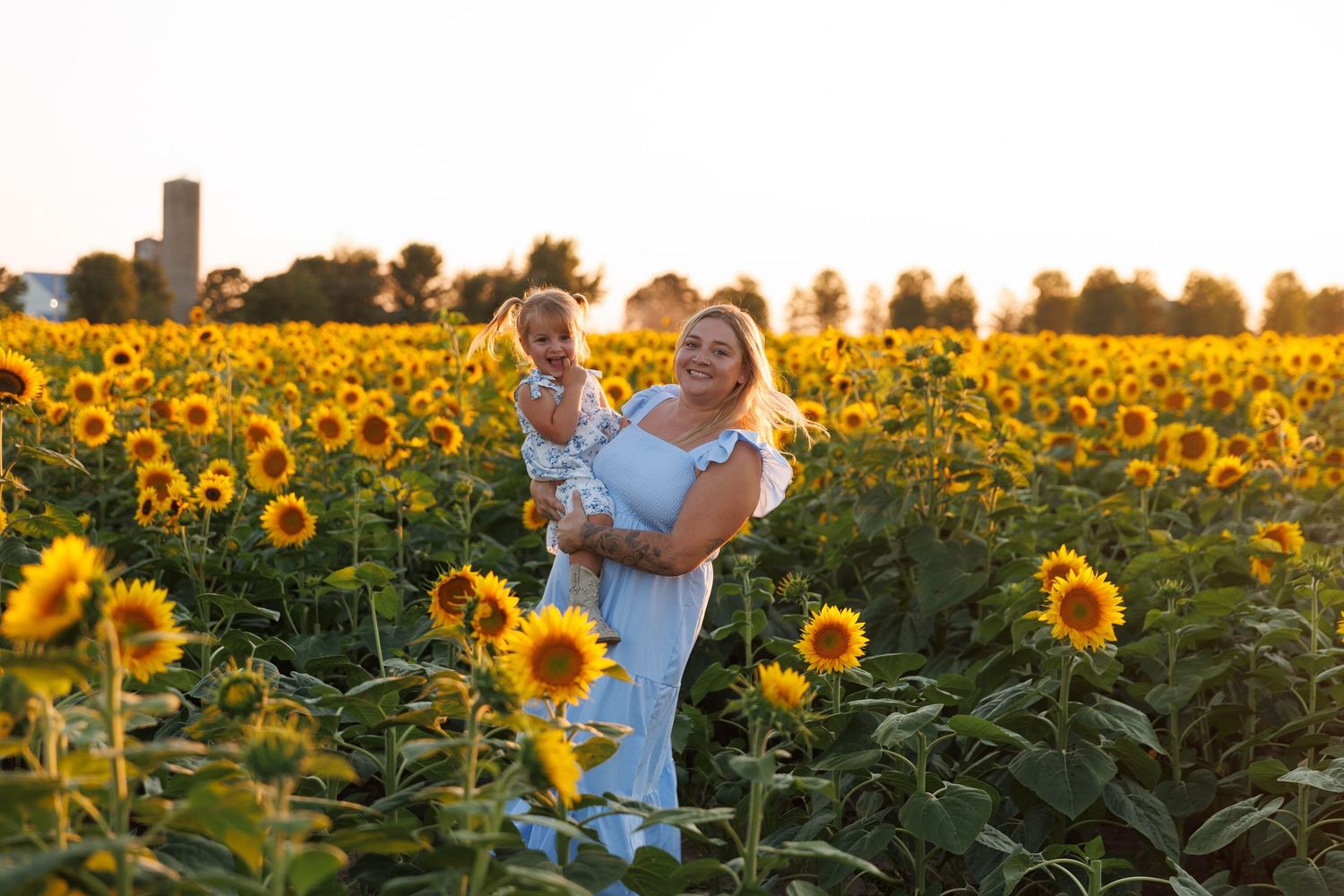 Mother holding toddler in white dresses standing in sunflower field at golden hour sunset.