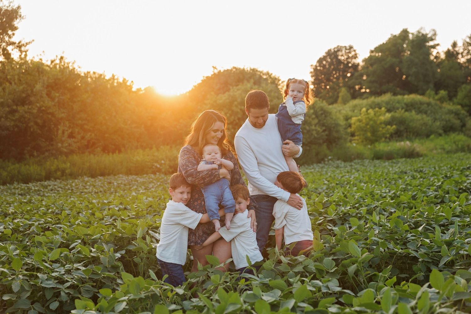 Loving family of six posing together in lush green farm field during warm sunset.