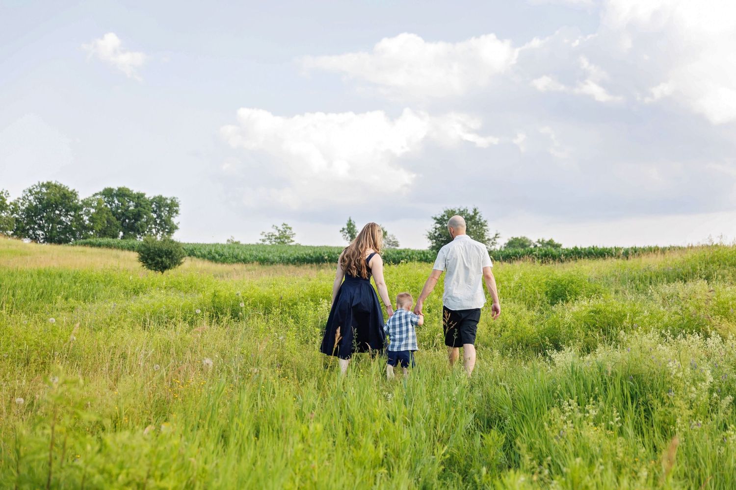 Family of three walking hand-in-hand through lush green field under dramatic cloudy sky.